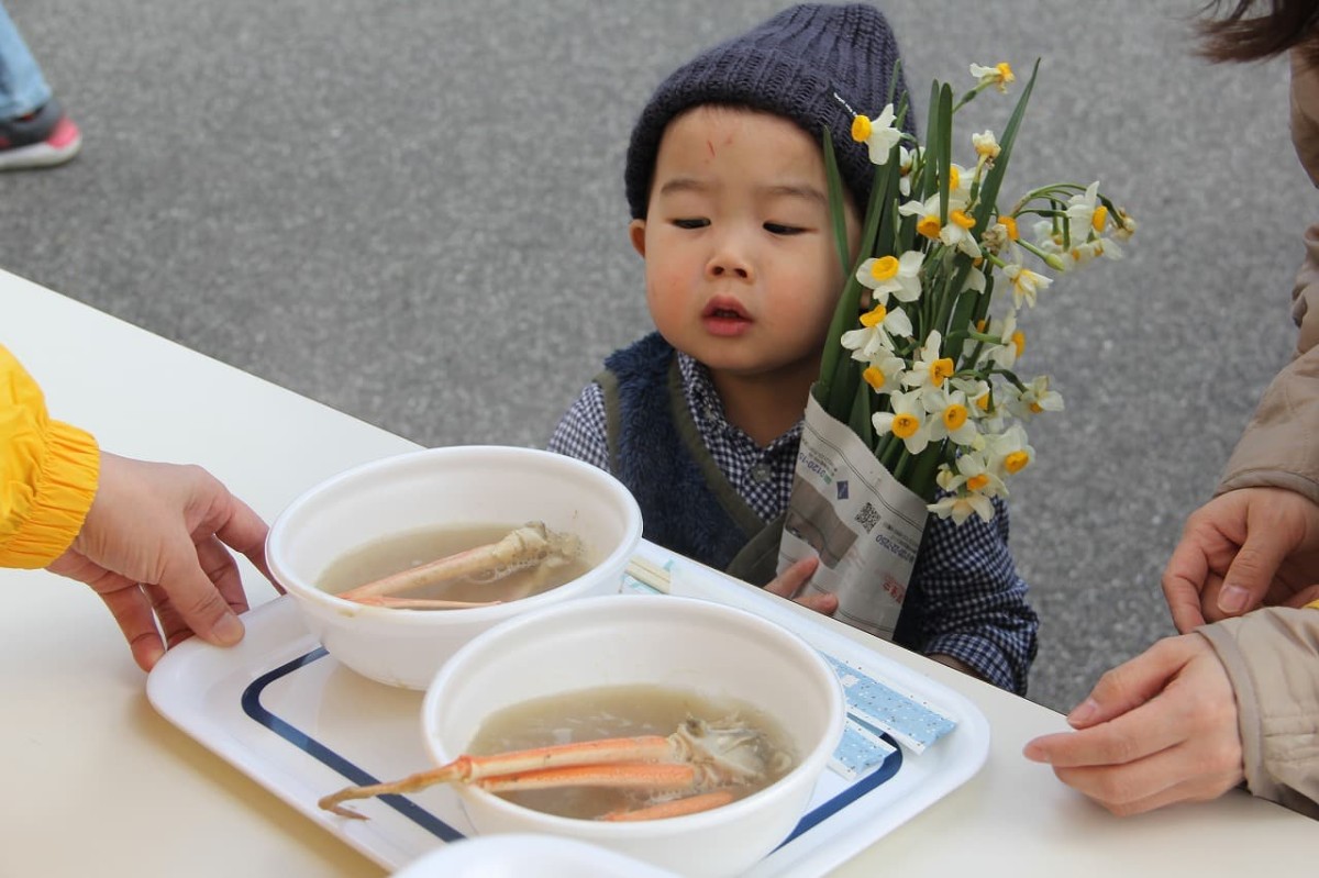 島根県隠岐郡で開催された『すいせん祭り』で配布したカニ汁と水仙を受け取る地元の子ども